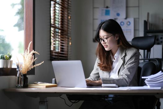 Asian Businesswoman Using laptop computer and working at office with calculator document on desk, doing planning analyzing the financial report, business plan investment, finance analysis concept..