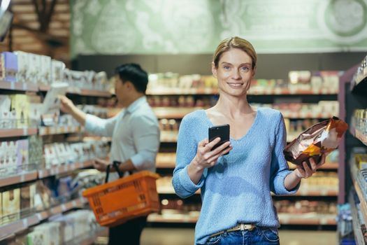 Portrait of a woman buyer in a supermarket in the grocery department, a blonde housewife chooses goods and makes purchases looking at the camera and smiling.