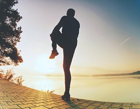 Athlete stretching lower body hamstring muscles before going running at lake in summer outdoors.