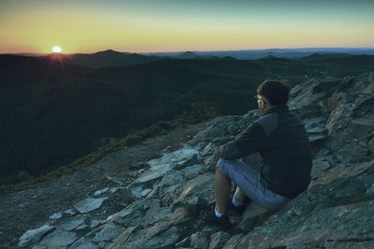 Man enjoying morning and looking to orange daybreak at misty horizon. Adult tourist in black sit on cliff's edge