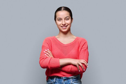 Portrait of teenage smiling female looking at camera on gray color studio background. Confident teen girl with crossed arms in red. Adolescence, high school, youth 15, 16 years old concept