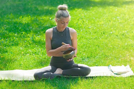 A slender woman in a gray top and leggings, sitting on the green grass in summer, holds a meditation bowl, drives a stick on it. Copy space