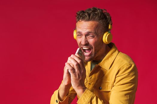 Handsome man 30s antics while pretend singing song using phone and wireless headphones wearing denim yellow jacket isolated on red background. Joyful man sing while listen music.