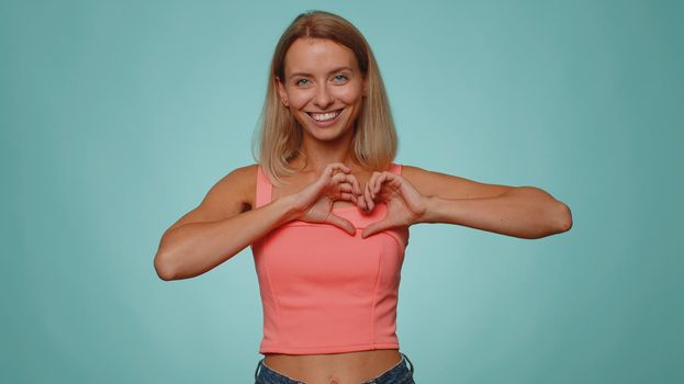 Woman in love. Smiling pretty woman 20s makes heart gesture demonstrates love sign expresses good feelings and sympathy. Young adult slim slender girl isolated alone on blue studio wall background