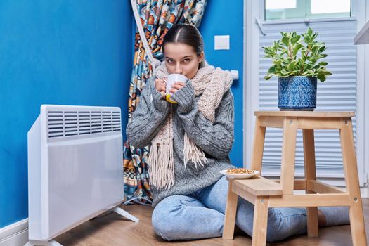 Cold autumn winter season, lifestyle, teenage female in warm sweater scarf warming near electric heating radiator, drinking hot drink with cup. Heating, warmth, equipment, life in cold season