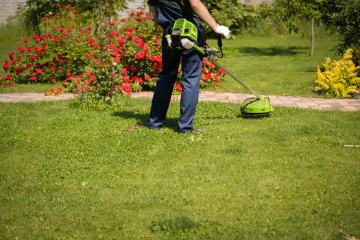 A young man is mowing a lawn with a lawn mower in his beautiful green floral summer garden. A professional gardener is cutting the grass in gloves.
