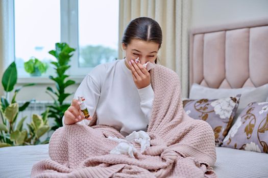 Young teenage female sitting in bed with runny nose medicine spray drops, sneezing into tissues, burying her nose. Autumn winter, rhinitis runny nose stuffy nose, treatment, flu season