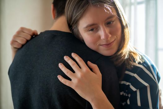 Happy young girl warmly hugs her boyfriend, rejoices at the meeting, stands near the window, smiley face in close-up, woman starts a romantic relationship. Husband and wife feel united, in a harmony.