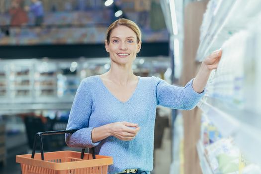 Portrait of a female buyer in a supermarket, a blonde housewife looking at the camera in the dairy department chooses milk and yogurt.