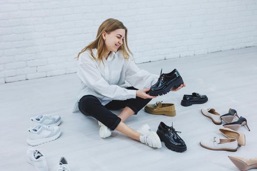 A woman sits on the floor in a dressing room between shoes and chooses new shoes for herself. Lady in a white shirt. Woman in white shoes.