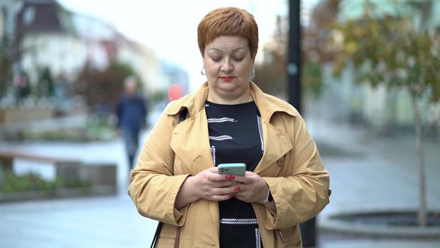 Serious woman with a phone in her hands on a city street. Middle-aged woman in autumn clothes standing and chatting online.