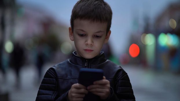 Portrait of a young boy with a phone in his hands is standing on the street in the evening. Child with a gadget at night against the background of blurred people and cars.