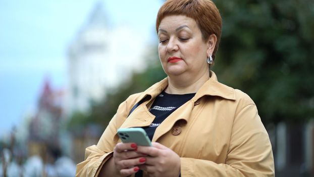 An elegant woman with short red hair and a raincoat is standing on the street and using a smartphone.