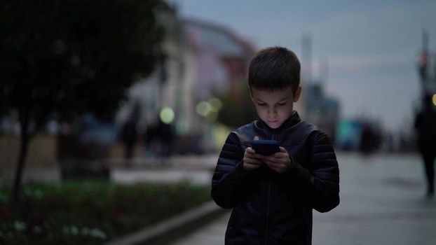Child boy with a modern smartphone walks through the night city along the street.