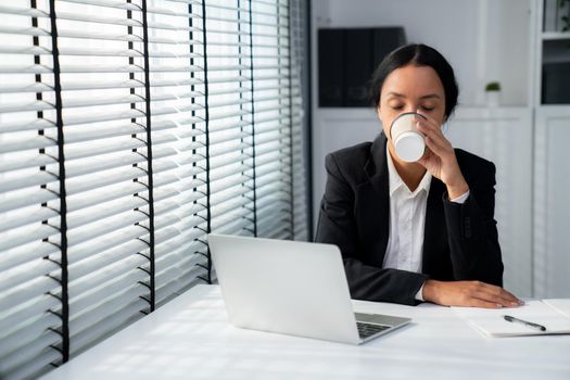 Competent female employee sits at her desk with a cup of coffee. Modern employee working with a drink, recreation during working hours, caffeine for people who are working