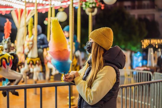 Young woman holding a cotton candy and smiling at a Christmas fair wearing a yellow wool cap wears a medical mask against coronavirus COVID 19.