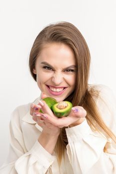 Portrait of a lovely smiling young brunette caucasian woman wearing the white shirt with long hair holding and showing avocado, standing isolated over white background