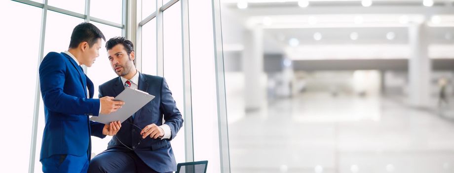 Two business people talk project strategy at office in widen view meeting room. Businessman discuss project planning with colleague at modern workplace .