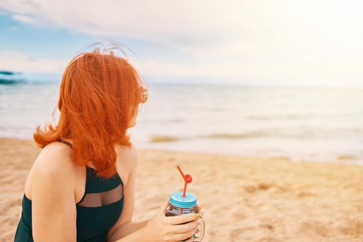 Cute girl in swimsuit is sitting on beach with cocktail and look at the ocean. Tropical alcohol beverage. Summer day with cloudy sky. Red-haired woman with sunglasses on vacation. Holidays at sea.