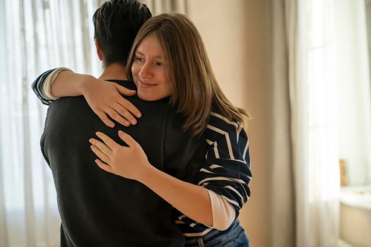 Happy young girl warmly hugs her boyfriend, rejoices at the meeting, stands near the window, smiley face in close-up, woman starts a romantic relationship. Husband and wife feel united, in a harmony.