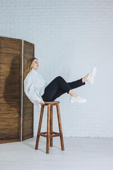 A beautiful woman is wearing a white cotton shirt and black pants. Cheerful emotional girl sits on a wooden chair against a white brick wall.