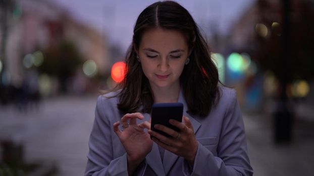 Portrait of a beautiful woman in a jacket using a smartphone on the street in the night city. Business lady stands with a phone on the background of blurred people and cars in the evening.