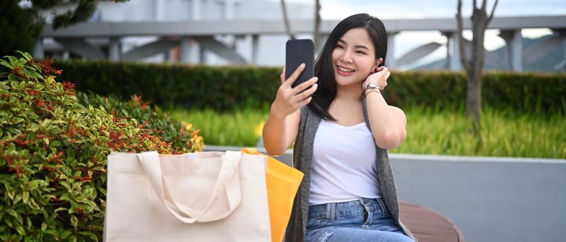 Cheerful young asian sitting on a park bench with shopping bags and takes a selfie on smart phone.