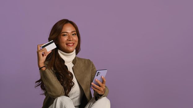 Studio photo of smiling young woman holding mobile phone and credit card sitting on purple background.