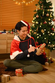 Man in Santa hat opening Christmas gifts and sitting in decorated room. Holidays and celebration concept.