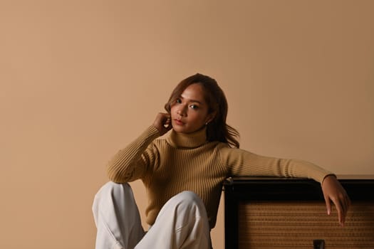 Portrait of fashionable woman wearing trendy sweater sitting near vintage radio, studio shot on beige background.