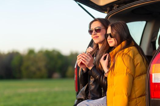 Mother and daughter camping on a hill and admiring the sunset while sitting in the car trunk