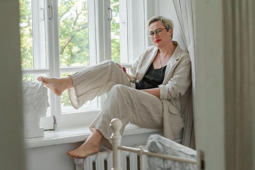 A middle-aged woman in a beige suit and black tank top sits mysteriously and looks out the window on the windowsill. Green trees outside.