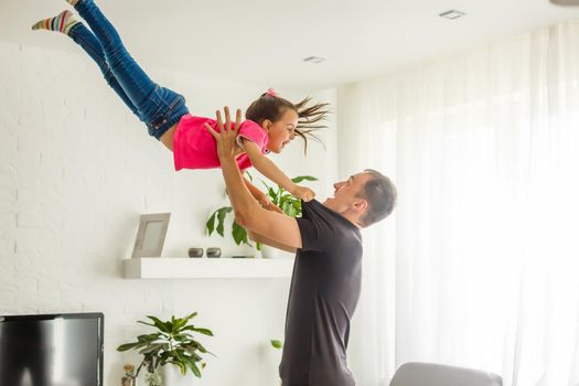 I can fly. Happy dad is lifting up his little daughter while standing in living room.