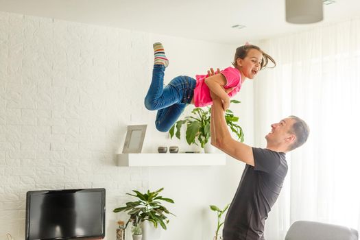 I can fly. Happy dad is lifting up his little daughter while standing in living room.