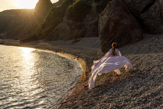 A mysterious female silhouette with long braids stands on the sea beach with mountain views, Sunset rays shine on a woman. Throws up a long white dress, a divine sunset