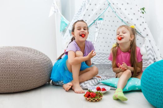 two little girls eat strawberries at home near wigwam tent