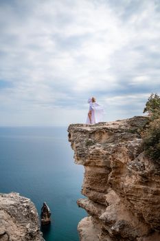 Blonde with long hair on a sunny seashore in a white flowing dress, rear view, silk fabric waving in the wind. Against the backdrop of the blue sky and mountains on the seashore