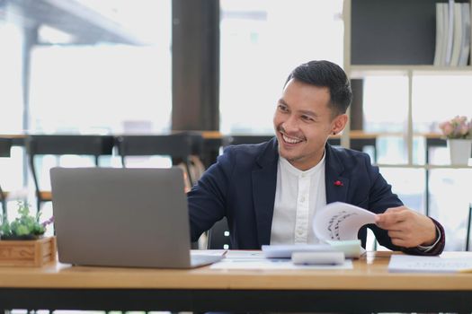 Portrait of a young Asian businessman smiling while using a laptop and writing down notes while sitting at his desk in a modern office.