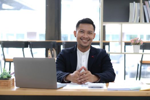 Portrait of a young Asian businessman smiling while using a laptop and writing down notes while sitting at his desk in a modern office.