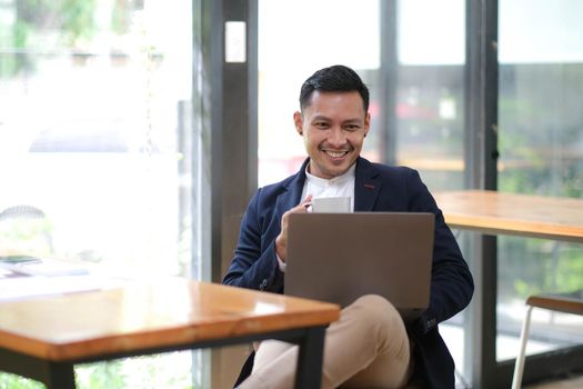 Young Asian businessman in suit and holding hot coffee cup and looking on laptop computer in office. Man in suit using laptop in well-lit workplace..