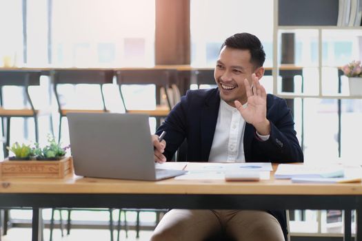 Portrait of a young Asian businessman smiling while using a laptop and writing down notes while sitting at his desk in a modern office.