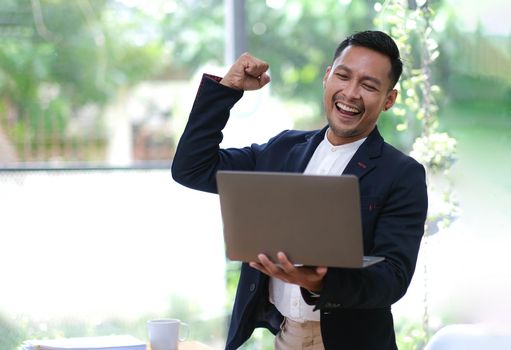 Asian businessman shouts with joy and holds hands up, celebrates victory, man works at desk in office, looks at laptop screen.