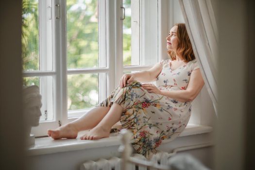 A middle-aged woman in a cream dress sits mysteriously and looks out the window on the windowsill. Green trees outside