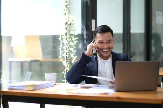 Asian businessman sitting on the phone with a customer with a laptop and document at his office desk..