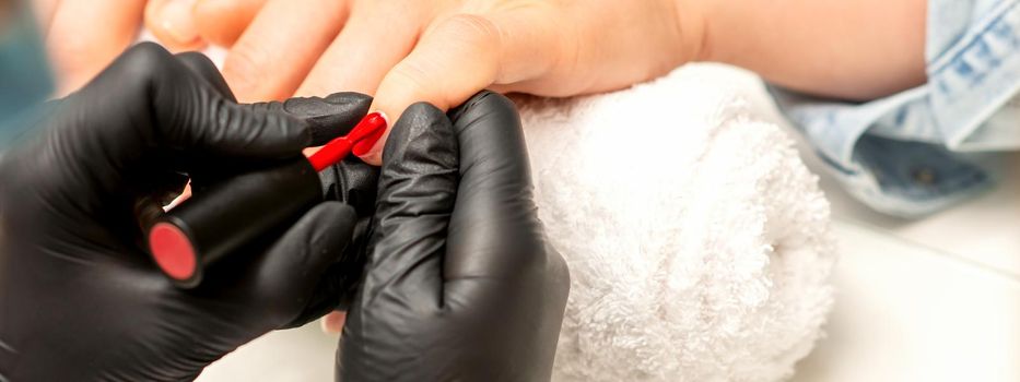 Manicure varnish painting. Close-up of a manicure master wearing rubber black gloves applying red varnish on a female fingernail in the beauty salon