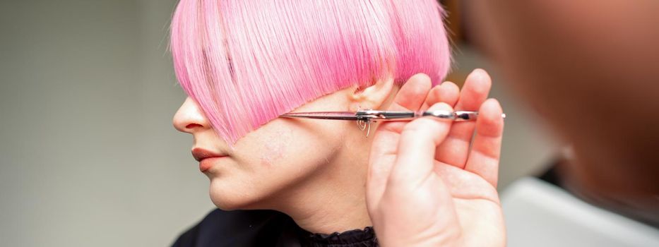 Hand of a hairdresser cutting short pink with scissor hair in a hairdressing salon, close up, side view