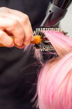 Drying short pink hair of young caucasian woman with a black hairdryer and black round brush by hands of a male hairdresser in a hair salon, close up