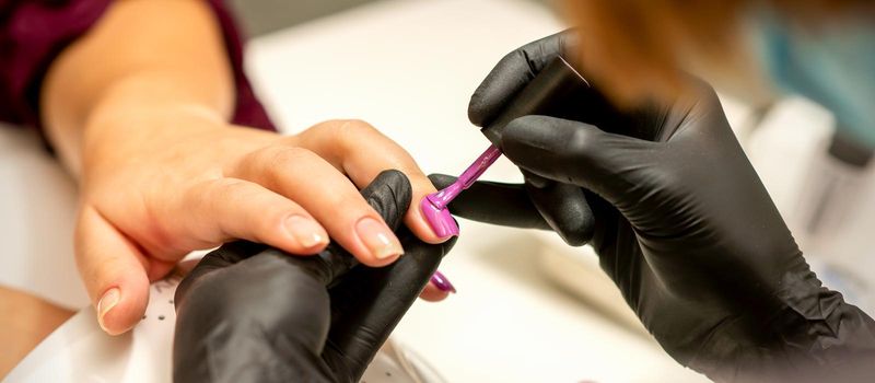 Professional manicure. A manicurist is painting the female nails of a client with purple nail polish in a beauty salon, close up. Beauty industry concept