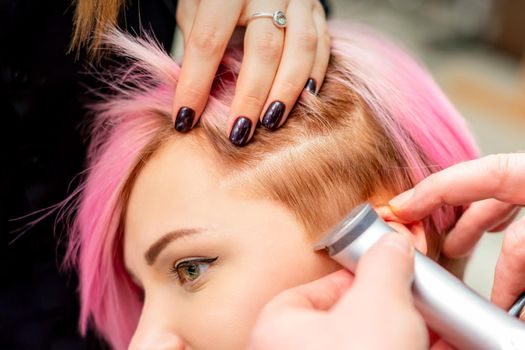 Hairdresser shaves female temple with pink hair by electric shaver in a hairdresser salon, close up