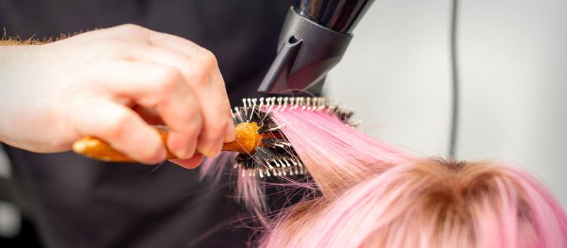 Drying short pink hair of young caucasian woman with a black hairdryer and black round brush by hands of a male hairdresser in a hair salon, close up